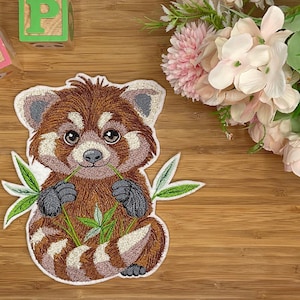 Könnte beinhalten: Gestickter Aufnäher eines roten Pandas mit braunem und weißem Fell, der grüne Bambusblätter hält. Der Aufnäher hat einen weißen Rand und ist vor einem Holzgrund platziert. Zwei Holzblöcke mit den Buchstaben "R" und "P" befinden sich im Hintergrund.