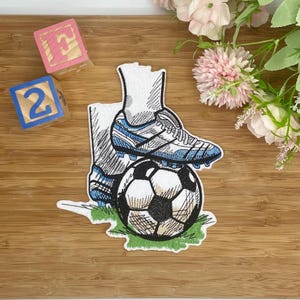 Könnte beinhalten: Ein gestickter Aufnäher mit einem Fußball und einem Fußballschuh mit blauen Akzenten, der auf einem Stück grünem Gras liegt. Der Aufnäher ist weiß mit schwarzen und blauen Details. Zwei Holzblöcke mit den Zahlen 2 und dem Buchstaben F befinden sich im Hintergrund.