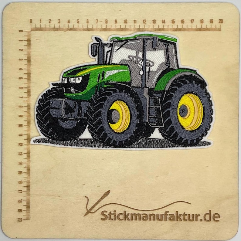 Puede incluir: Parche bordado de un tractor verde y gris con ruedas amarillas. El tractor est&aacute; detallado con neum&aacute;ticos negros y una cabina gris. El parche est&aacute; sobre una superficie de madera con una regla y el texto "Stickmanufaktur.de".