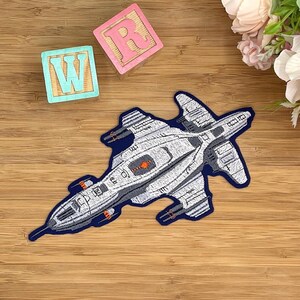 Könnte beinhalten: Gestickter Aufnäher eines futuristischen Raumschiffs in Grau- und Weißtönen, umrandet in Marineblau. Der Aufnäher weist detaillierte Designelemente und orangefarbene Akzente auf. Der Aufnäher befindet sich auf einer Holzoberfläche mit Spielzeugblöcken und Blumen.