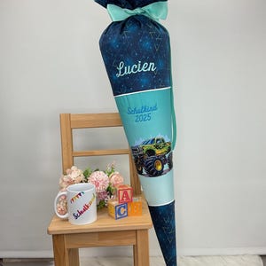 Könnte beinhalten: Eine große, blau-türkisfarbene Schultüte mit dem Namen "Lucien" darauf gestickt. Die Tüte hat ein geometrisches Muster und eine Monster-Truck-Grafik. Ein passender Becher und ABC-Blöcke stehen auf einem kleinen Holzstuhl.