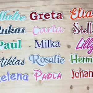 Puede incluir: Parches bordados con nombres variados, como Malte, Greta y Elias, en diferentes colores y fuentes. Cada parche tiene un borde blanco. Presentados sobre una superficie de madera clara.