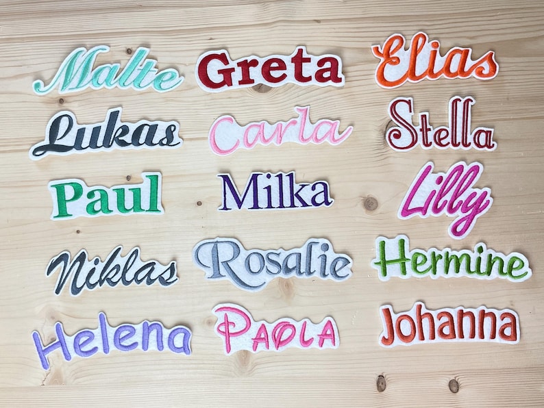 Puede incluir: Parches bordados con nombres variados, como Malte, Greta y Elias, en diferentes colores y fuentes. Cada parche tiene un borde blanco. Presentados sobre una superficie de madera clara.