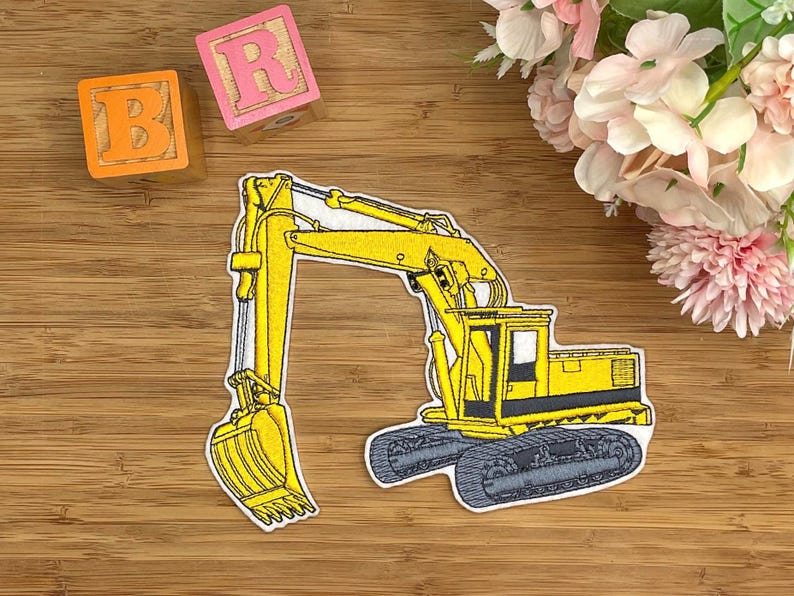 K&ouml;nnte beinhalten: Ein gelber, gestickter Aufn&auml;her eines Bau-Baggers mit schwarzen und grauen Ketten. Der Bagger hat eine gro&szlig;e Schaufel und ein detailliertes Design. Zwei Holzbl&ouml;cke mit den Buchstaben "B" und "R" sind sichtbar.