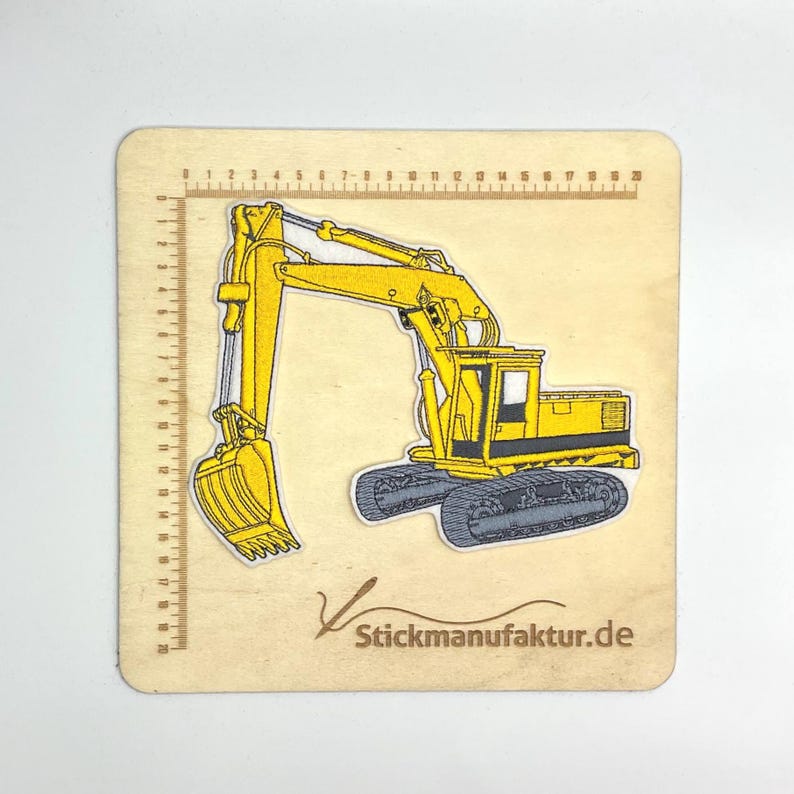 K&ouml;nnte beinhalten: Ein gelber Bagger-Aufn&auml;her mit schwarzen Ketten und detailliertem Design. Der Aufn&auml;her befindet sich auf einem Holzquadrat mit einem aufgedruckten Lineal an den R&auml;ndern. Der Text "Stickmanufaktur.de" ist unten rechts aufgedruckt.