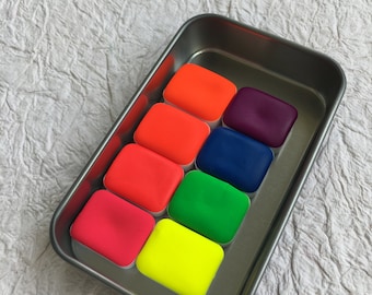May include: A metal tin containing eight watercolour paint pans. The pans are filled with bright neon colours, including pink, orange, red, yellow, green, blue, purple, and black.