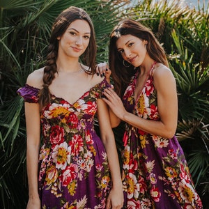 May include: Two women wearing floral print dresses. The dresses are a deep purple with a vibrant pattern of red, yellow, and white flowers. One dress has off-the-shoulder sleeves, while the other has a halter neckline. The background features lush green foliage and a body of water.