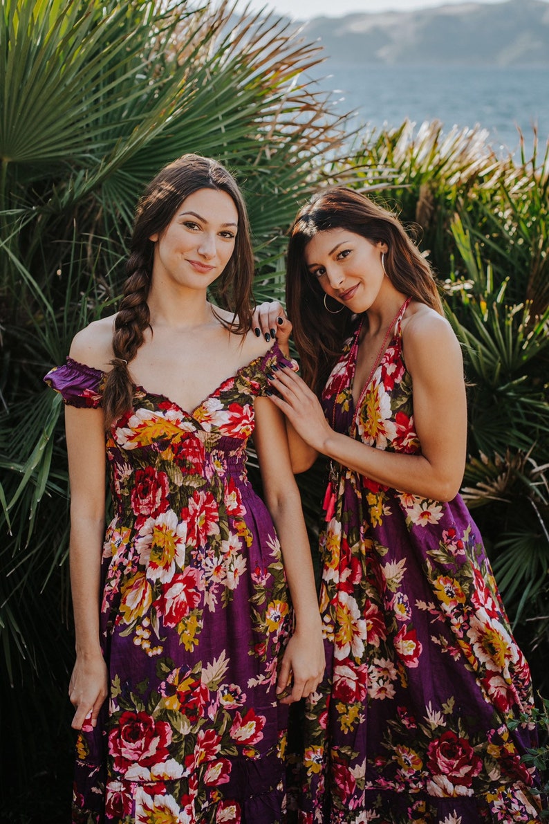 May include: Two women wearing floral print dresses. The dresses are a deep purple with a vibrant pattern of red, yellow, and white flowers. One dress has off-the-shoulder sleeves, while the other has a halter neckline. The background features lush green foliage and a body of water.