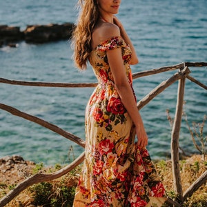 May include: A woman wearing a long, off-the-shoulder floral dress with red and yellow roses. The dress has a flowing skirt. She is standing near a wooden fence with the ocean and mountains in the background.