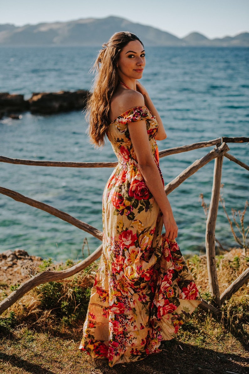 May include: A woman wearing a long, off-the-shoulder floral dress with red and yellow roses. The dress has a flowing skirt. She is standing near a wooden fence with the ocean and mountains in the background.