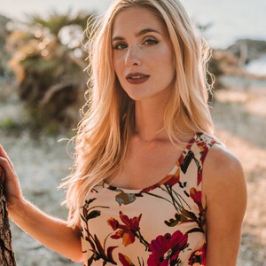 May include: A woman with long blonde hair is wearing a sleeveless floral dress with a white background and red, yellow, and green flowers. She is standing in front of a tree and looking at the camera.