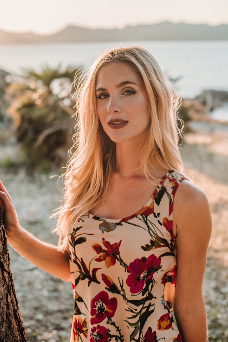 May include: A woman with long blonde hair is wearing a sleeveless floral dress with a white background and red, yellow, and green flowers. She is standing in front of a tree and looking at the camera.