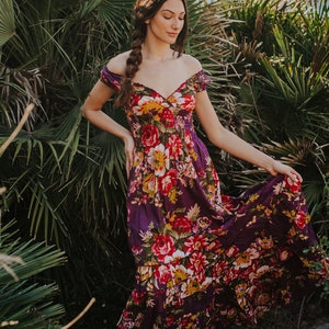 May include: A long, flowing dress with an off-the-shoulder neckline. The dress features a vibrant floral pattern in shades of red, yellow, and white against a deep purple background. The model is standing outdoors near lush green foliage.