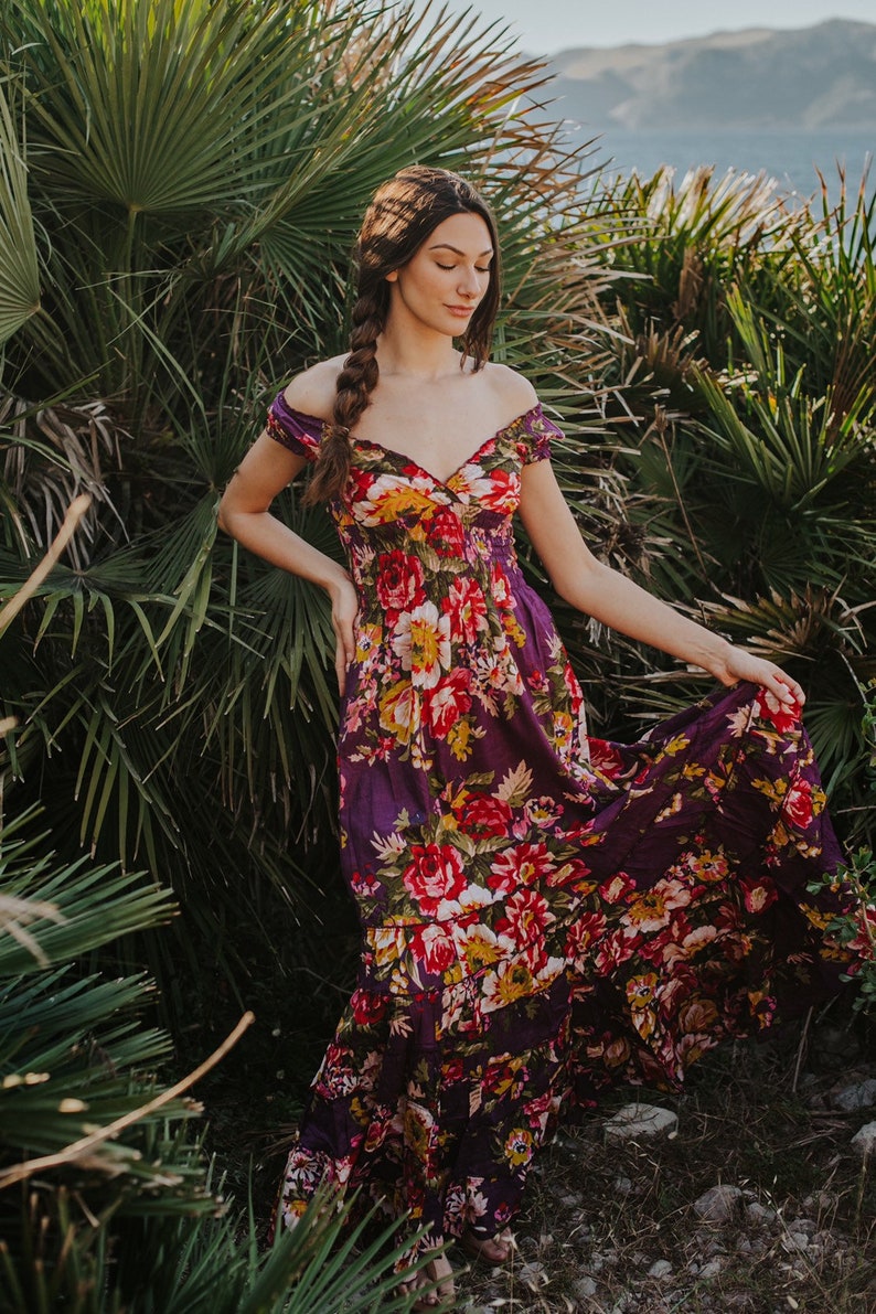 May include: A long, flowing dress with an off-the-shoulder neckline. The dress features a vibrant floral pattern in shades of red, yellow, and white against a deep purple background. The model is standing outdoors near lush green foliage.