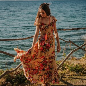 May include: A flowing, off-the-shoulder maxi dress with a floral pattern in shades of red, orange, and yellow. The dress is worn outdoors near a body of water, with a wooden fence in the foreground. A long, orange tassel necklace is visible.