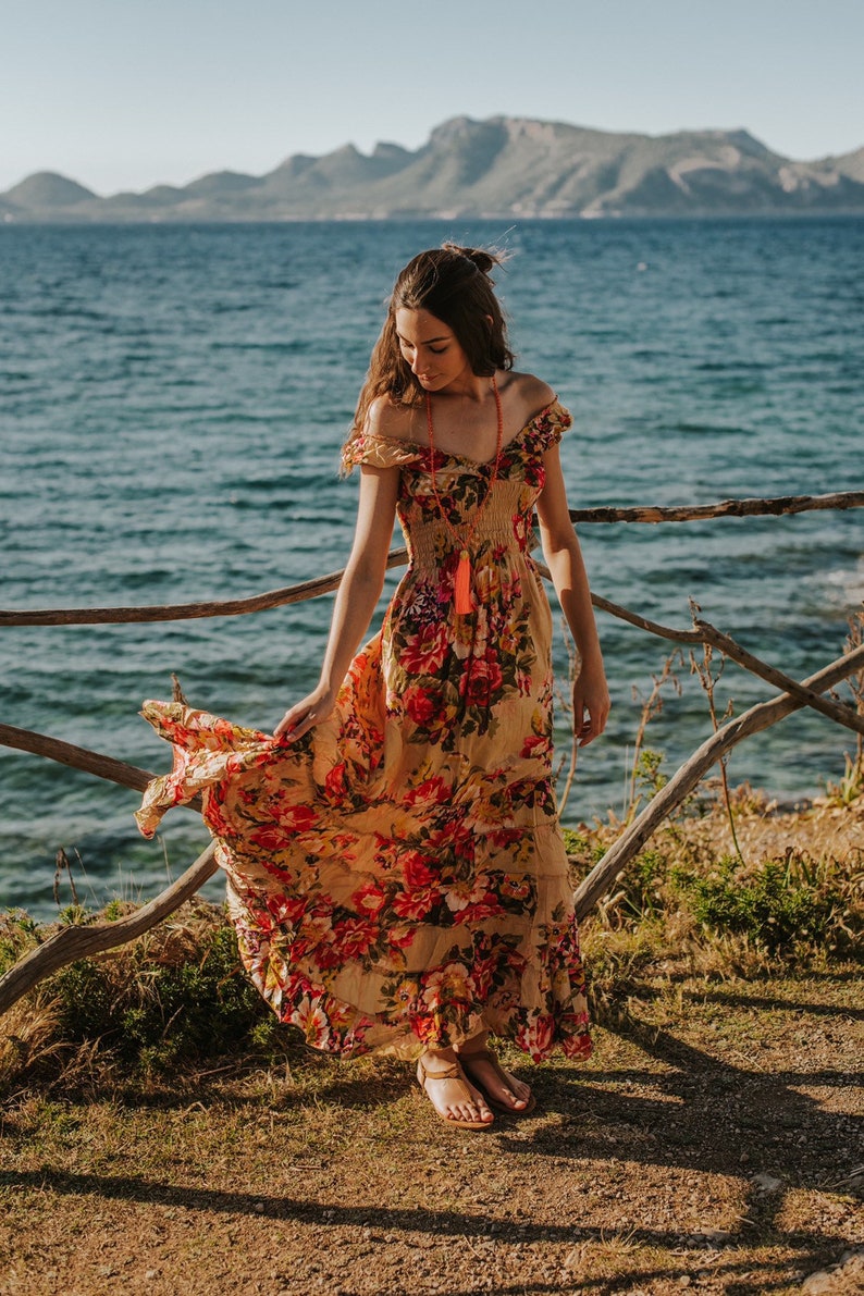 May include: A flowing, off-the-shoulder maxi dress with a floral pattern in shades of red, orange, and yellow. The dress is worn outdoors near a body of water, with a wooden fence in the foreground. A long, orange tassel necklace is visible.