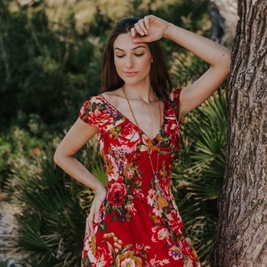 May include: A red floral print dress with a V-neck and short sleeves. The dress features a vibrant pattern of large roses and other flowers in shades of pink, yellow, and green. A long beaded necklace with a tassel hangs down the front.
