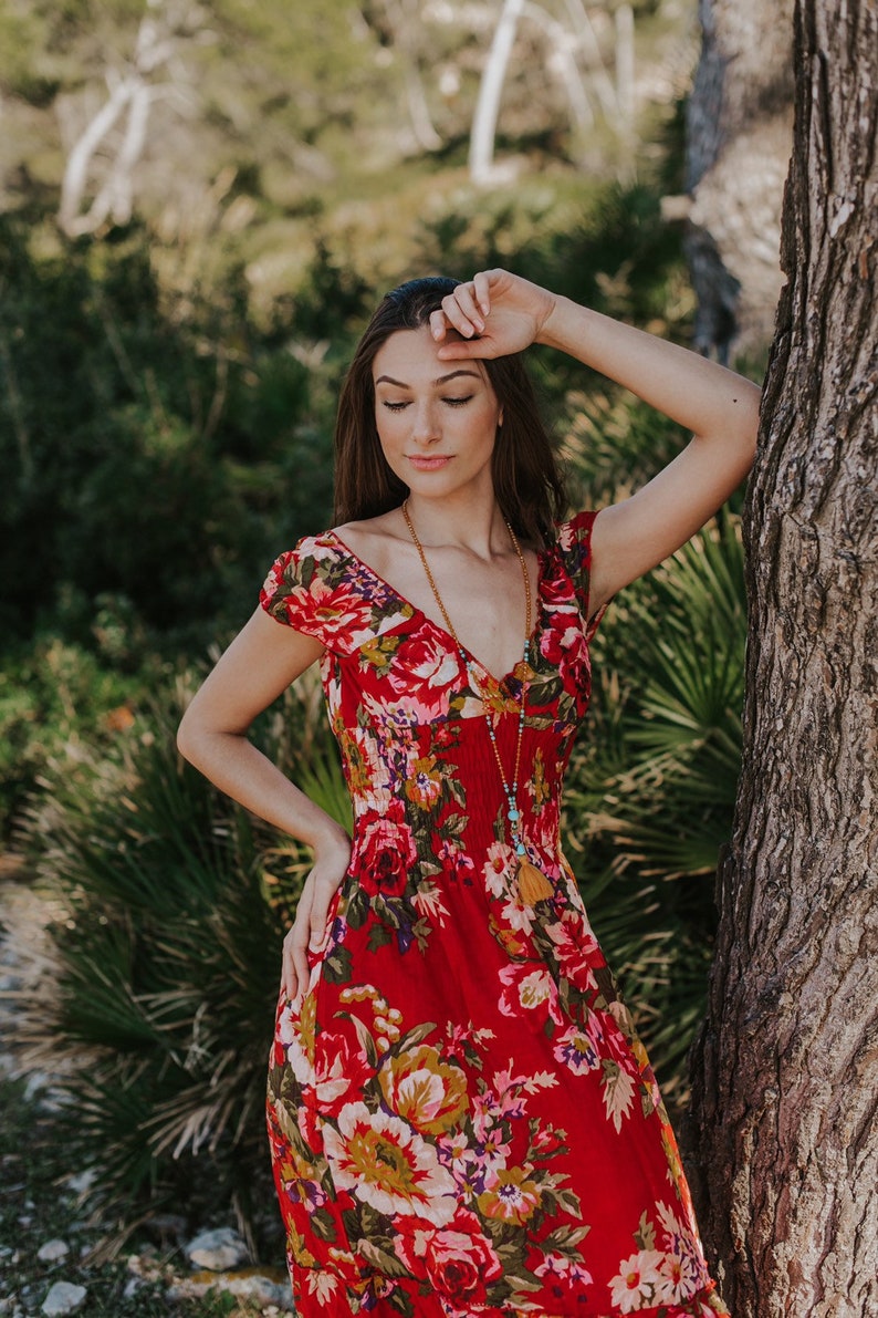 May include: A red floral print dress with a V-neck and short sleeves. The dress features a vibrant pattern of large roses and other flowers in shades of pink, yellow, and green. A long beaded necklace with a tassel hangs down the front.