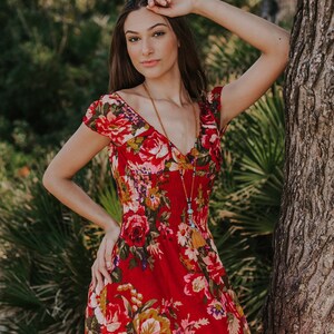 May include: A red floral print dress with a V-neck and short sleeves. The dress features a vibrant pattern of red roses, yellow and green foliage. A long beaded necklace with a tassel is also visible.
