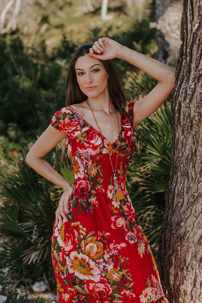 May include: A red floral print dress with a V-neck and short sleeves. The dress features a vibrant pattern of red roses, yellow and green foliage. A long beaded necklace with a tassel is also visible.