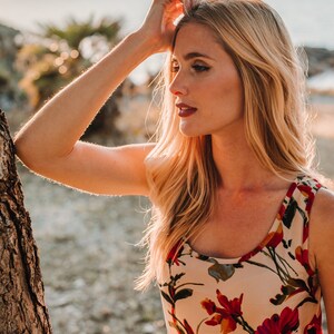 May include: A woman with long blonde hair wearing a sleeveless floral top with red, yellow, and green flowers on a cream background. She is standing outdoors with a tree trunk to her left.