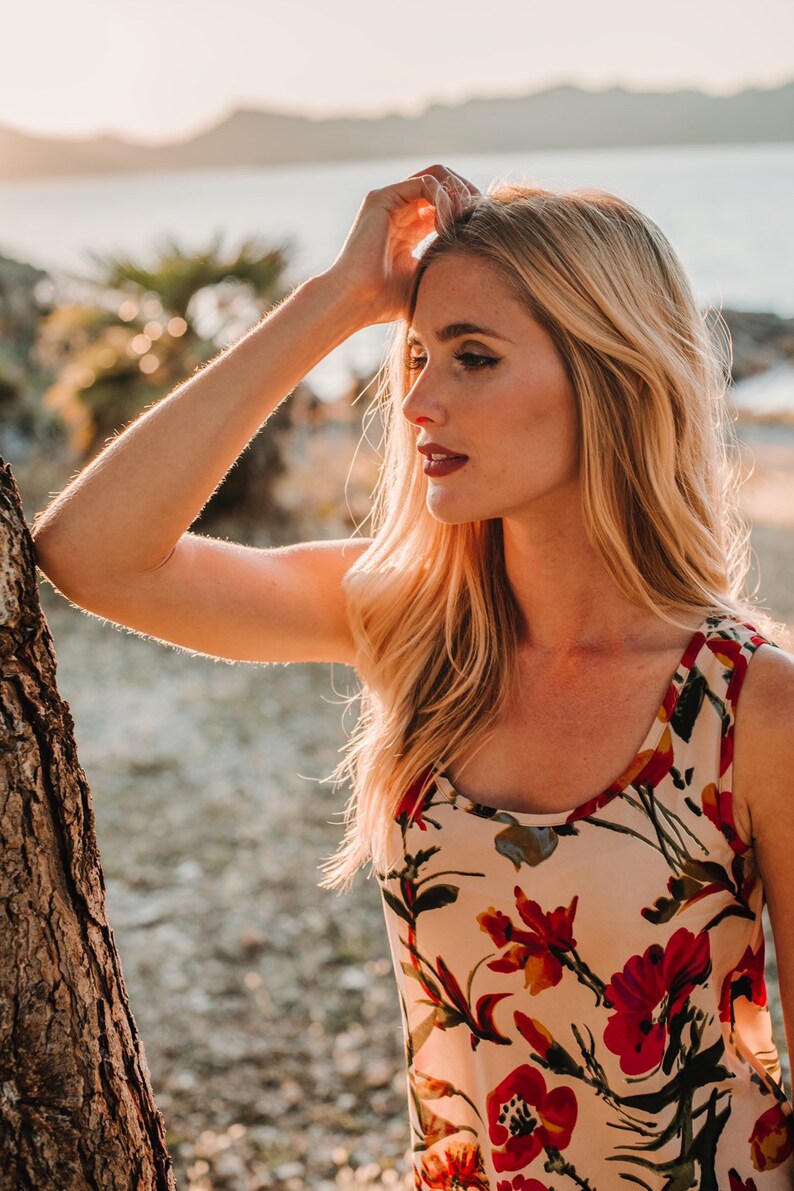 May include: A woman with long blonde hair wearing a sleeveless floral top with red, yellow, and green flowers on a cream background. She is standing outdoors with a tree trunk to her left.