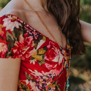 May include: A red floral print dress with a scoop neckline. A long gold beaded necklace with turquoise accents and a tassel hangs over the dress. The dress has a fitted bodice and short sleeves.