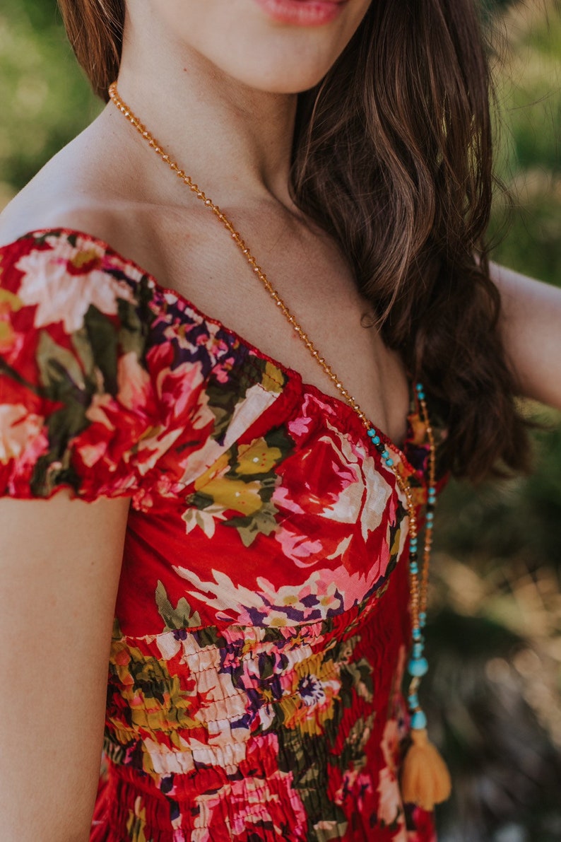 May include: A red floral print dress with a scoop neckline. A long gold beaded necklace with turquoise accents and a tassel hangs over the dress. The dress has a fitted bodice and short sleeves.