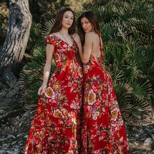 May include: Two women wearing red floral maxi dresses. The dresses feature a vibrant floral pattern with shades of pink, yellow, and white. One dress has short sleeves and a V-neck, while the other has a halter-style back. The dresses are floor-length.