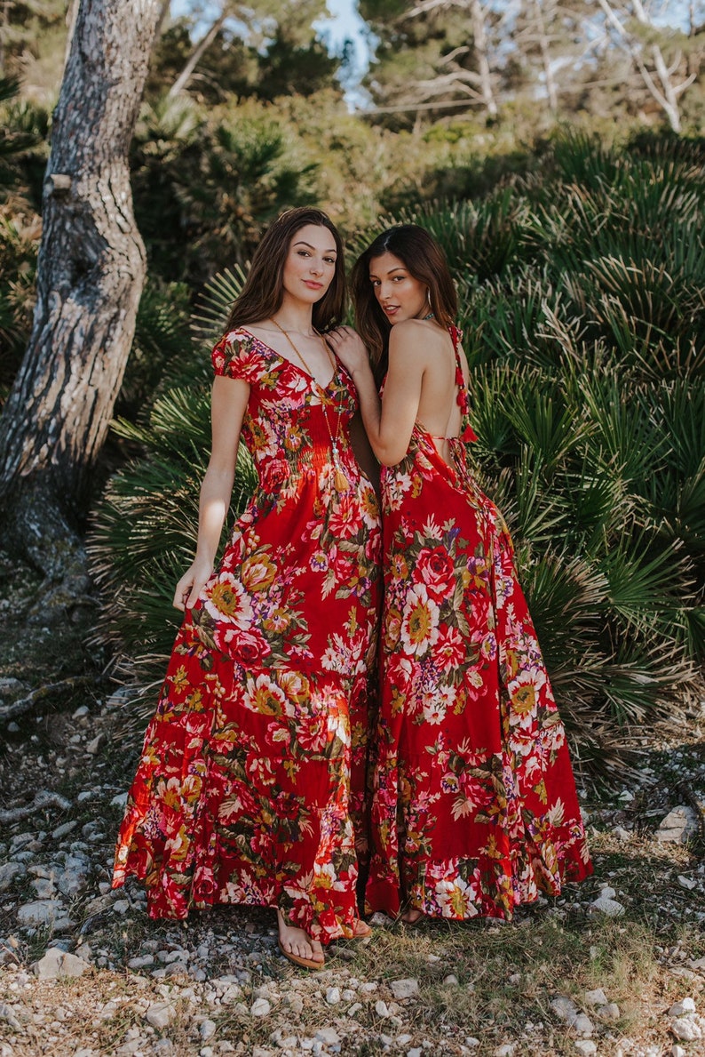 May include: Two women wearing red floral maxi dresses. The dresses feature a vibrant floral pattern with shades of pink, yellow, and white. One dress has short sleeves and a V-neck, while the other has a halter-style back. The dresses are floor-length.