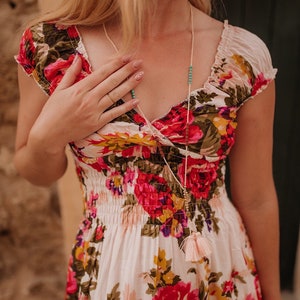 May include: A floral print dress with a V-neckline and short sleeves. The dress features a pattern of red, pink, and yellow roses on a white background. A long necklace with turquoise beads and a pink tassel is also visible.