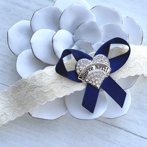 May include: A white lace garter with a navy blue ribbon and a silver heart charm that says "You're Next!" The charm is decorated with rhinestones.