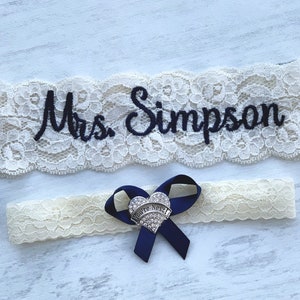 May include: White lace garter with blue ribbon and a heart-shaped charm that says "You're Next". The garter is embroidered with the text "Mrs. Simpson".