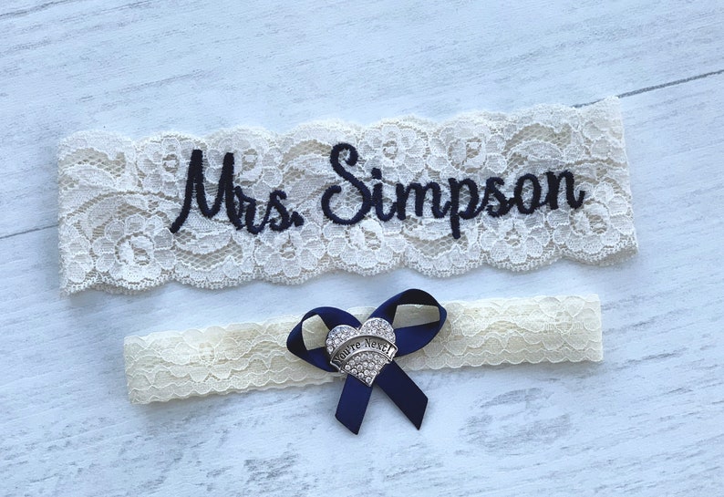 May include: White lace garter with blue ribbon and a heart-shaped charm that says "You're Next". The garter is embroidered with the text "Mrs. Simpson".