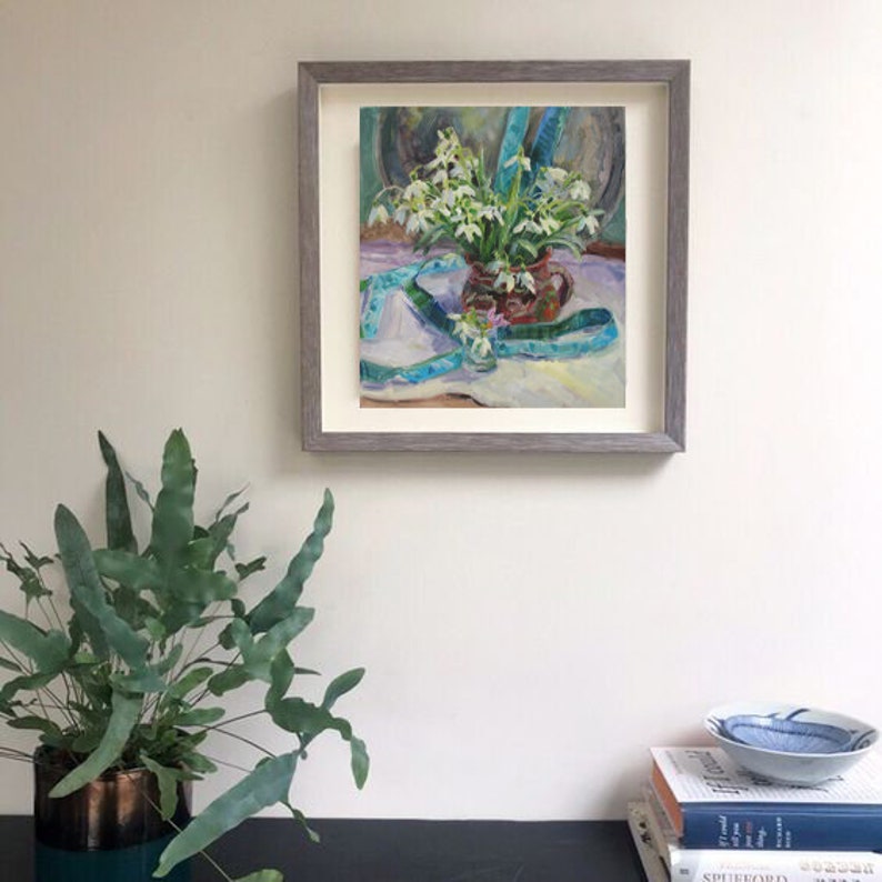 May include: A framed painting of white flowers in a brown vase with a blue ribbon. The painting is in a gray frame and is hanging on a white wall. A potted plant is in the foreground.