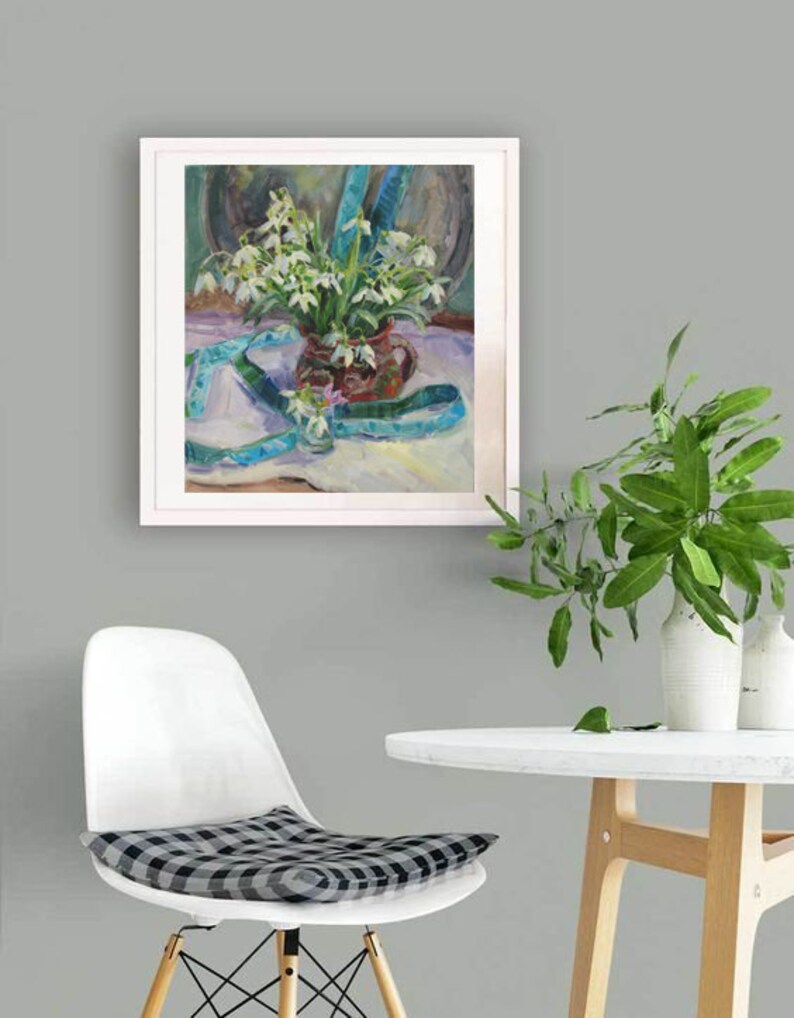 May include: A framed oil painting depicts a still life of white flowers in a brown vase, with a blue ribbon. The painting is set against a neutral background. A white chair with a checkered cushion and a small white table are in the foreground.