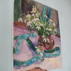 May include: Oil painting of a still life featuring a bouquet of white snowdrop flowers in a red vase. A turquoise ribbon and a small glass with snowdrops are also present. The painting has a soft, impressionistic style with a light color palette.
