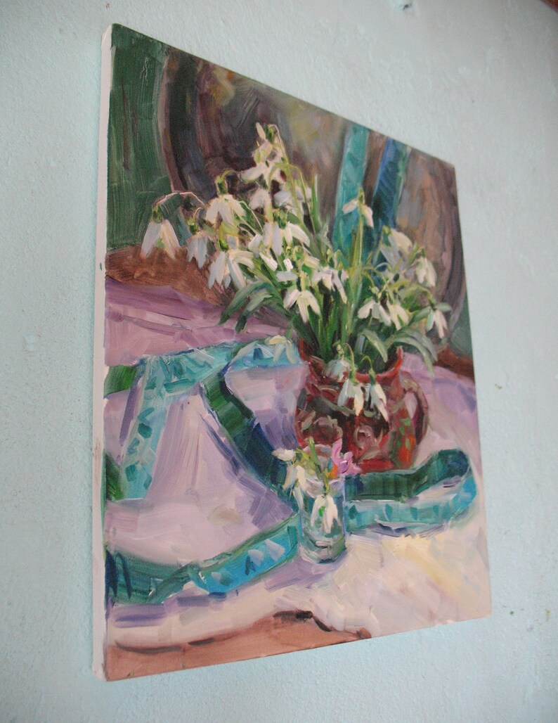 May include: Oil painting of a still life featuring a bouquet of white snowdrop flowers in a red vase. A turquoise ribbon and a small glass with snowdrops are also present. The painting has a soft, impressionistic style with a light color palette.