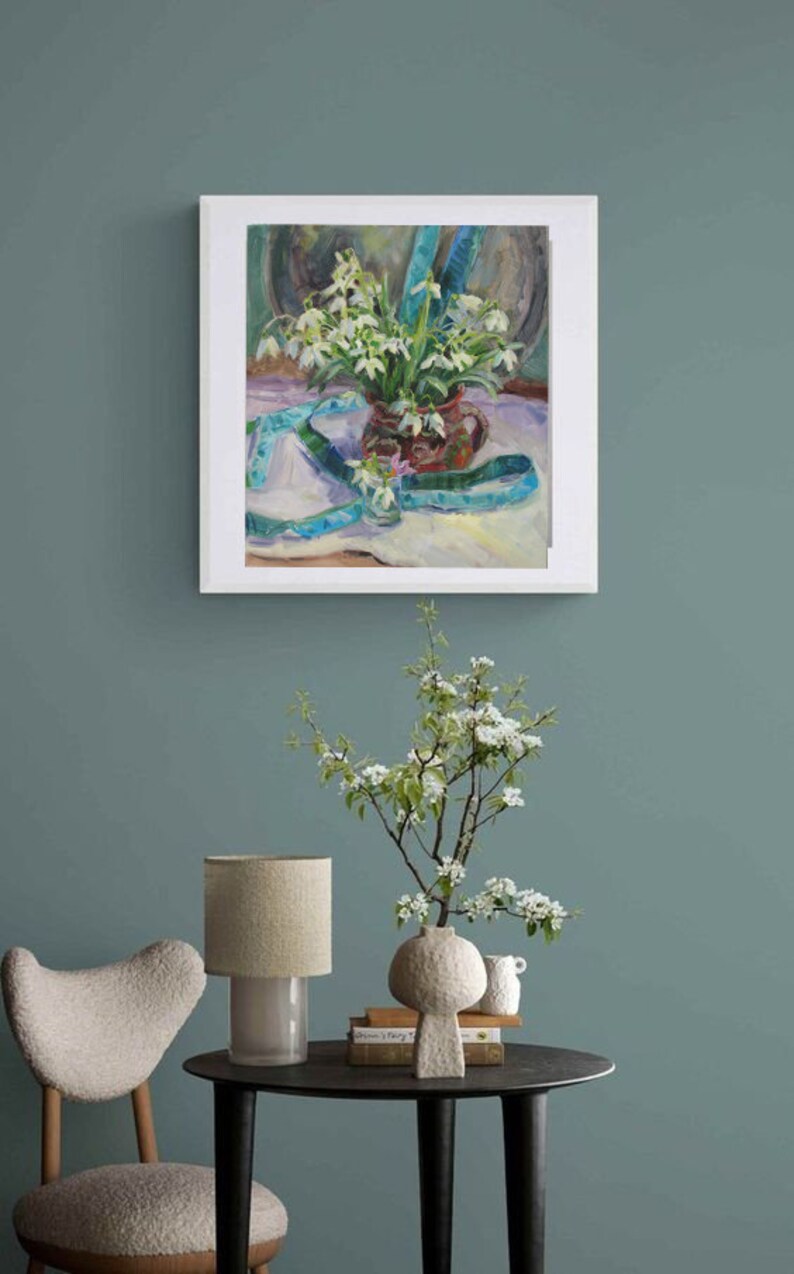 May include: An oil painting of a still life featuring a bouquet of white flowers in a brown vase, accented with a blue ribbon. The painting is framed and hangs on a teal wall. A side table holds a lamp, vase, and books.