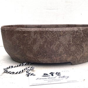 May include: A brown, oval planter with a textured surface. The planter has a low profile and sits on three small feet. A tag with black and white string is attached to the planter.