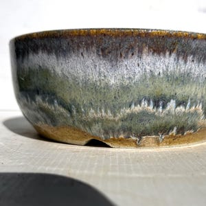 May include: A handcrafted ceramic bowl with a distinctive glaze. The bowl showcases a mix of green, white, and brown hues, producing a textured, natural look. The bowl is round with a gently sloping rim, suitable for serving.