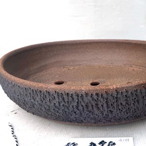 May include: Oval-shaped brown ceramic bonsai pot with a textured, dark brown exterior. The pot has two drainage holes and a smooth interior. The rim has a rough, bark-like texture. A perfect container for bonsai trees.