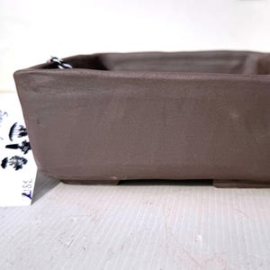 May include: A brown, rectangular ceramic bonsai pot with a textured surface. The pot has a low profile and sits on four small feet. The pot is designed for growing small trees and plants. The tag says "Summerland Pottery".