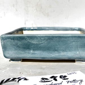 May include: A rectangular, teal ceramic bonsai pot with a textured surface. The pot has a square shape and sits on four small feet. The interior is white. A tag with black and white string is in the foreground.