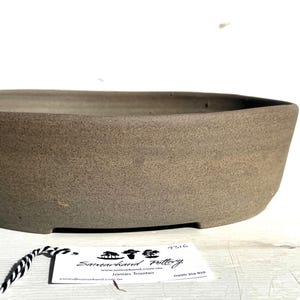 May include: A brown, oval ceramic bonsai pot with a textured surface. The pot has a low profile and sits on small feet. A tag with text is attached to the pot with a black and white string. The pot is designed for bonsai trees.