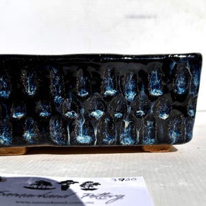 May include: Rectangular ceramic planter with a textured, dark blue glaze. The surface has a pattern of raised, rounded shapes with blue and white accents. The planter sits on small, tan feet.