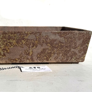 May include: Rectangular brown planter with a textured surface and a mottled pattern of green and brown. The planter has a slightly angled design and is suitable for indoor or outdoor use.