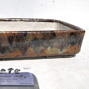 May include: A rectangular brown and green glazed ceramic bonsai pot with a white interior. The pot has a label with the text "Samarkand Pottery" and the number "3487".
