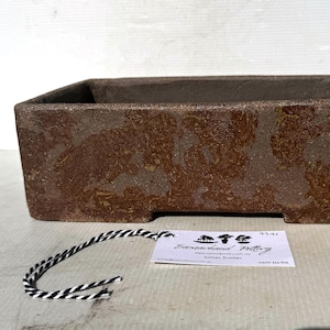 May include: A rectangular brown ceramic bonsai pot with a textured, mottled glaze finish. A small white rectangular tag with black text reading Samarkand Pottery and a black and white striped string are positioned in front of the pot.