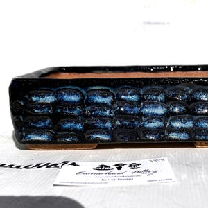 May include: A rectangular bonsai pot with a textured, dark blue exterior and a brown interior. The pot has a raised, brick-like pattern on the sides and a rectangular shape. The pot is made of ceramic and has a glossy finish.